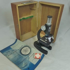 May include: A classic black microscope with a silver eyepiece and stage, beside an open wooden case. A blue booklet titled "world within a world" and a small box of "COVER SLIPS" are also visible.