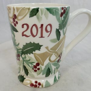 Puede incluir: Taza de cerámica blanca con asa, con el año "2019" en borgoña. La taza está decorada con hojas verdes de acebo, bayas rojas y adornos dorados en forma de trompeta, ideal para las fiestas.