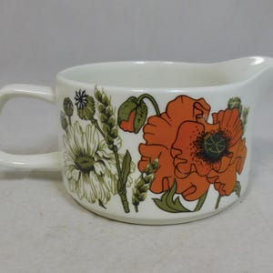 May include: White ceramic creamer with a handle and spout, decorated with orange poppies, green leaves, and other floral elements. The creamer has a curved handle and a spout for easy pouring. The floral design is in shades of orange, green, and white.