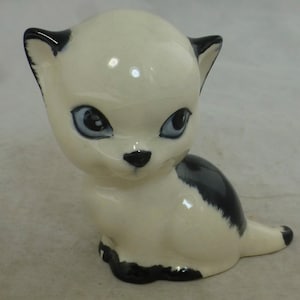 May include: A white ceramic figurine of a cat with black markings. The cat has large, blue eyes and a black nose.