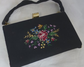 以下が含まれることがあります： 黒いハンドバッグ、花の刺繍デザイン。 刺繍された花は赤、青、黄色、紫の色合いです。 バッグは金色の留め金と黒いハンドルが付いています。