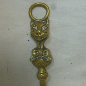 Könnte beinhalten: Ein Kaminwerkzeug aus Messing mit einem Katzenkopfdesign. Die Katze hat ein weißes Gesicht und eine schwarze Schleife. Das Werkzeug hat oben einen Ring und die Worte "Cheshire Cat" sind unten eingraviert.