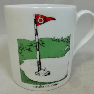 Peut inclure: Une tasse en céramique blanche avec une scène de terrain de golf vert, blanc et rouge. La tasse a une anse et dit "mole in one" au fond.