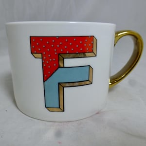 May include: White ceramic mug with a gold handle. The mug features a red and blue 3D letter "F" with white polka dots on the red section.