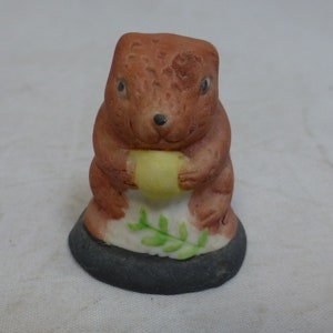May include: A small ceramic figurine of a brown bear holding a yellow ball. The bear is sitting on a base with green leaves.