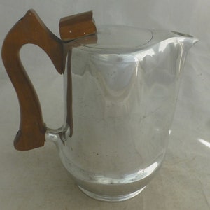 Puede incluir: Una cafetera de plata con un mango de madera. La cafetera tiene un cuerpo redondeado y un pico en el lateral. El mango es curvo y tiene un pomo de madera en la parte superior.