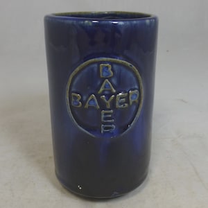 May include: A dark blue ceramic mug with a raised, circular design featuring the word "Bayer" in a gold-coloured font.