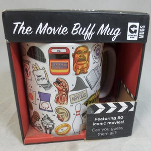 Op de afbeelding: Een witte keramische mok met verschillende filmthema-illustraties, verpakt in een zwarte doos. De mok toont afbeeldingen van een typemachine, een hersen, een haaienvin en meer. De doos leest "The Movie Buff Mug" en "Featuring 50 iconic movies!"