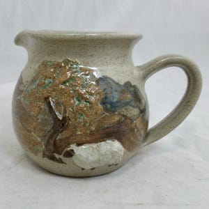 Puede incluir: Una pequeña jarra de cerámica de color crema con un asa marrón. La jarra tiene un diseño rústico pintado a mano con un árbol, una oveja y un paisaje rocoso.