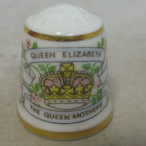 May include: White porcelain thimble with a gold trim and a floral design. The thimble features a crown and the text "Queen Elizabeth The Queen Mother's".