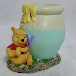 May include: A ceramic Winnie the Pooh honey pot figurine. The pot is painted in shades of yellow, white, and blue. Winnie the Pooh is sitting next to the pot, holding his paws together.