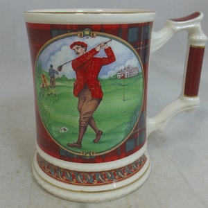 May include: A white ceramic mug with a red, green, and blue plaid design. The mug features a golfer swinging a club on a green golf course with a white building in the background. The mug has a gold rim and a gold handle.