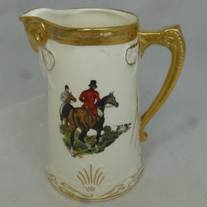 Peut inclure: Un pichet en céramique blanche avec un bord et une poignée dorés. Le pichet présente une scène peinte de deux personnes à cheval dans un champ.