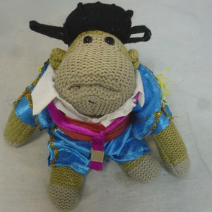 May include: A knitted monkey toy wearing a blue and pink matador costume with a black hat. The monkey has a white shirt and gold trim on the costume.