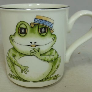 Könnte beinhalten: Weiße Keramiktasse mit einem grünen Frosch, der einen blauen und gelben Hut trägt. Der Frosch lächelt und hat seine Hände vor sich gefaltet.