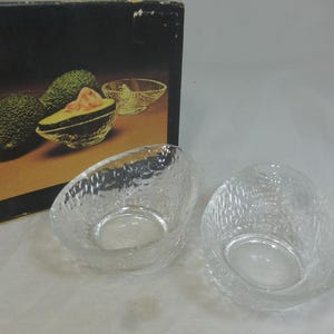 May include: Two clear, textured glass bowls in an avocado shape. The bowls have a textured surface and a rounded base. The bowls are next to a box with an avocado illustration.