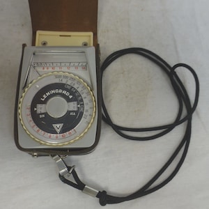 May include: A silver and black light meter with a leather case and a black strap. The light meter has a dial with the text "LENINGRAD 4" and numbers from 2 to 14. The case has a small flap that can be closed to protect the light meter.