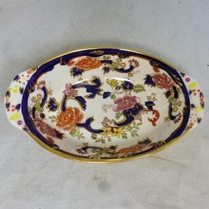 Op de afbeelding: Ovale keramische serveerschaal met een witte basis, met een bloemenpatroon in oranje, roze en geel. De schaal heeft een gouden rand en een donkerblauwe rand met decoratieve krullen. Handvatten zijn aan elk uiteinde aanwezig.