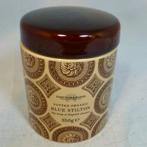 May include: A cylindrical ceramic jar with a dark brown lid and a cream-colored body. The body is decorated with a repeating pattern of floral and geometric designs in brown. Text on the jar reads "Potted Organic Blue Stilton" and "350g".