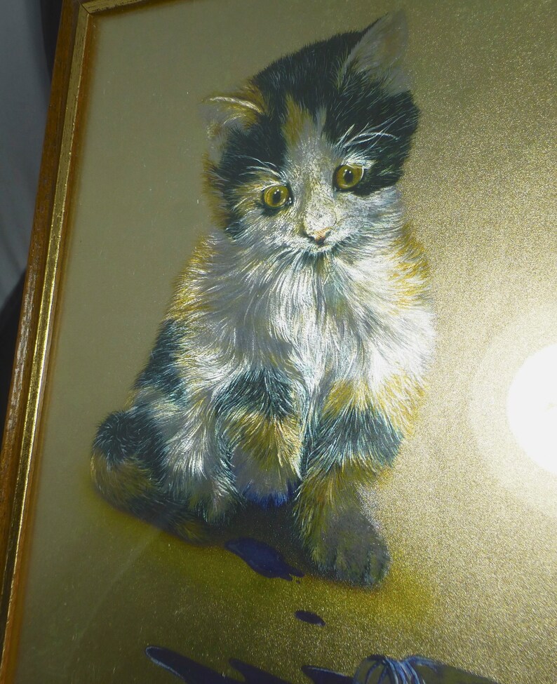Puede incluir: Una pintura enmarcada de un gatito calico con pelaje negro, blanco y naranja. El gatito est&aacute; sentado sobre un fondo dorado con una sombra azul y negra debajo.