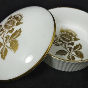 以下が含まれることがあります： 金色の縁取りと花柄の白い磁器製の小物入れ。蓋には金色の葉のついたバラが1輪、箱には花柄が繰り返し描かれている。