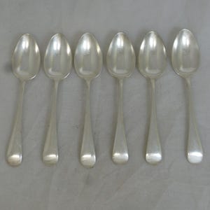 Puede incluir: Seis cucharas plateadas dispuestas en fila. Cada cuchara tiene un cuenco redondeado y un mango largo y delgado. Las cucharas son de un metal brillante y reflectante, probablemente acero inoxidable o plata. Las cucharas son para comer.