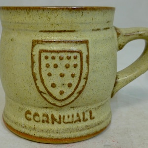 May include: A light green ceramic mug with a brown handle and a brown crest with the word "Cornwall" printed below.