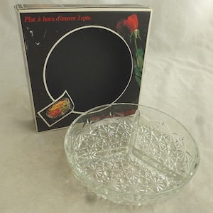 Puede incluir: Un plato de servir de vidrio transparente con tres secciones divididas. El plato tiene un patrón decorativo de pequeñas estrellas. El plato está en su caja original, que es negra con una rosa roja en la parte delantera. La caja dice "Plat à hors d'œuvre 3 cpts."