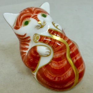 Puede incluir: Figura de gato de cerámica con rayas rojas y blancas y detalles dorados. El gato está en una pose juguetona, con ojos verdes y una sonrisa. Las orejas y las patas son blancas.