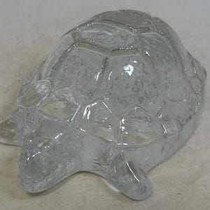 May include: A clear glass turtle figurine with a textured surface. The turtle has a detailed shell pattern and visible legs and head. The figurine is a decorative item.