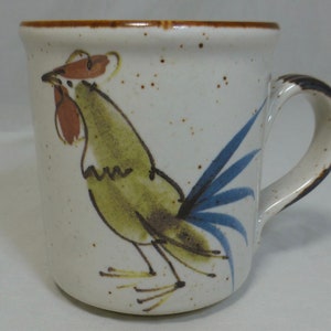 以下が含まれることがあります： 茶色の縁と取っ手のある白いセラミック製のマグカップ。マグカップには、緑と茶色の羽根と青い尾を持つ雄鶏の手描きデザインが施されています。