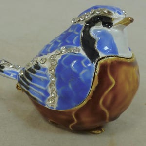 May include: A decorative bird figurine in blue, brown, black, and white, embellished with rhinestones. The bird has a hinged lid and is likely a trinket box. The figurine is made of a glossy material and has gold-coloured accents.