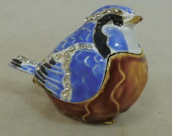 May include: A decorative bird figurine in blue, brown, black, and white, embellished with rhinestones. The bird has a hinged lid and is likely a trinket box. The figurine is made of a glossy material and has gold-colored accents.