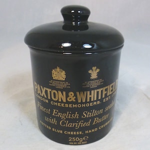以下が含まれることがあります： 金色の蓋と金色の文字が書かれた黒い陶器の壺。壺には「Paxton & Whitfield London Cheesemongers Est. 1797」と「Finest English Stilton sealed with Clarified Butter Potted Blue Cheese, Hand Crumbled 250g」と書かれている。