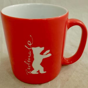 Puede incluir: Una taza de cerámica de color rojo brillante con interior blanco y un asa en el lado derecho. El exterior presenta un gráfico blanco de la silueta de un oso de pie junto al texto vertical blanco que dice Berlinale en letra cursiva.