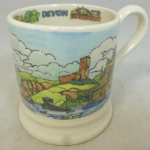 May include: A cream-colored ceramic mug featuring a colorful illustration of a coastal landscape with a stone castle, boats on the water, and green hills. The interior rim displays the text DEVON in blue letters alongside small tea-themed graphics.