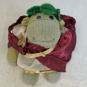 May include: A knitted plush toy in the shape of a sheep, dressed in a burgundy cape and a green laurel wreath. The sheep has a light green body, gray feet, and a white and gold-trimmed garment. A gold-colored button adorns the cape.