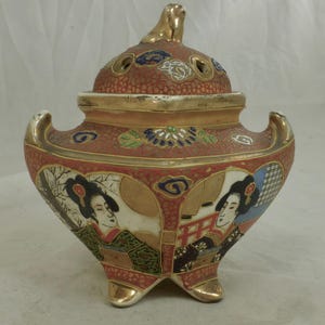 May include: A vintage, ornate ceramic incense burner with a lid. The red body features gold accents and painted panels depicting geishas. The lid has air holes and a gold finial. The burner rests on three gold feet.