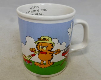 May include: White ceramic mug with a cartoon image of Garfield the cat mowing the lawn and grilling. The mug has the text "Happy Father's Day, Big Deal." printed on the front.