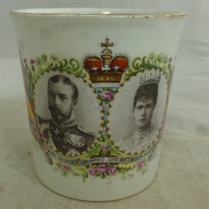 Könnte beinhalten: Weiße Keramiktasse mit floralem Design und Bildern von König Georg V. und Königin Mary. Der Text lautet "King George V Crowned June 1911 Queen Mary".