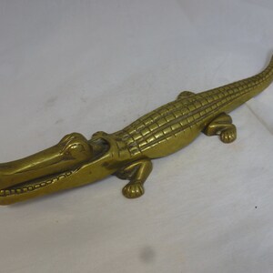 May include: A brass alligator nutcracker with a detailed, textured body and open mouth. The nutcracker is in a resting position with its legs extended.