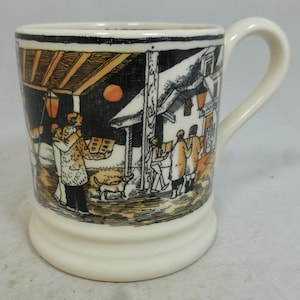 Puede incluir: Taza de cerámica color crema con una ilustración detallada de una escena invernal. La obra de arte representa figuras con abrigos, un perro y edificios con faroles. La taza tiene un asa curva y una base ligeramente acampanada.