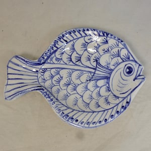 Puede incluir: Plato de cerámica blanco con forma de pez con detalles azules pintados a mano. El plato presenta un diseño detallado de escamas, aletas y una boca muy abierta. La combinación de colores azul y blanco le da un toque náutico.