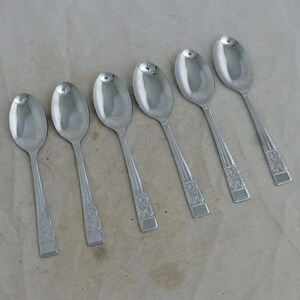 May include: A set of six silver-toned metal spoons arranged in a diagonal row. Each spoon features a smooth, oval-shaped bowl and a handle decorated with a subtle, embossed floral pattern near the base, ending in a flat, squared-off tip.