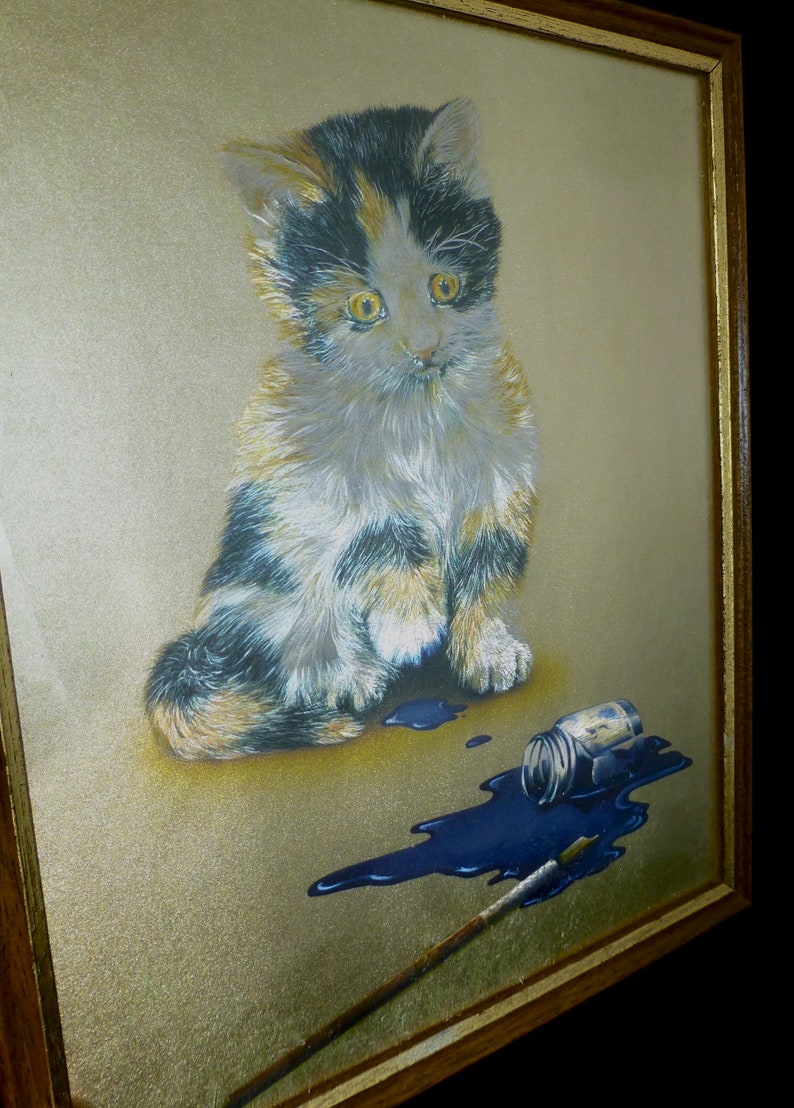 Puede incluir: Una pintura enmarcada de un gatito calico sentado sobre un fondo dorado con un frasco de pintura azul derramado y un pincel.