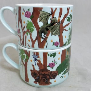 May include: Two stacked ceramic mugs with light blue exteriors and white interiors. Each mug features a whimsical forest scene with a dragon, a three-headed dog, and a fairy. The design includes brown trees, pink roses, and green foliage.