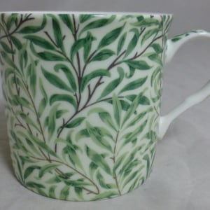 Könnte beinhalten: Eine weiße Keramiktasse mit einem grün-weißen Blättermuster. Die Tasse hat einen gebogenen Henkel und eine zylindrische Form. Die Blätter sind in verschiedenen Grüntönen dargestellt und erzeugen ein natürliches, botanisches Design. Die Tasse ist für Heißgetränke geeignet.