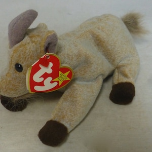 Puede incluir: Un juguete de peluche marrón claro con nariz y pezuñas marrones. El juguete tiene un cuerno morado y una cola marrón. El juguete está acostado de lado con la cabeza girada hacia la izquierda. El juguete tiene una etiqueta Ty roja unida a su cuello con el texto "Beanie Babies" y una estrella.