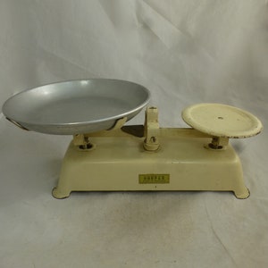 Op de afbeelding: Een vintage witte keukenweegschaal met twee metalen kommen. De weegschaal heeft een geel label met de tekst "HARPER MADE IN ENGLAND".