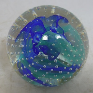 May include: A clear glass paperweight with blue and teal swirls. The interior design features a flowing pattern of colours, with small air bubbles throughout the glass. A decorative and artistic object.